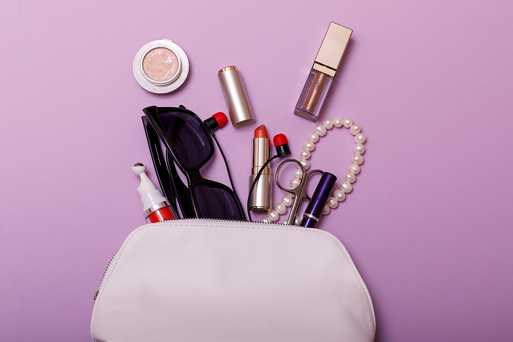Make up bag with cosmetics isolated on purpule background. Overhead view of essential beauty items sunglasses earphones and red rouge.