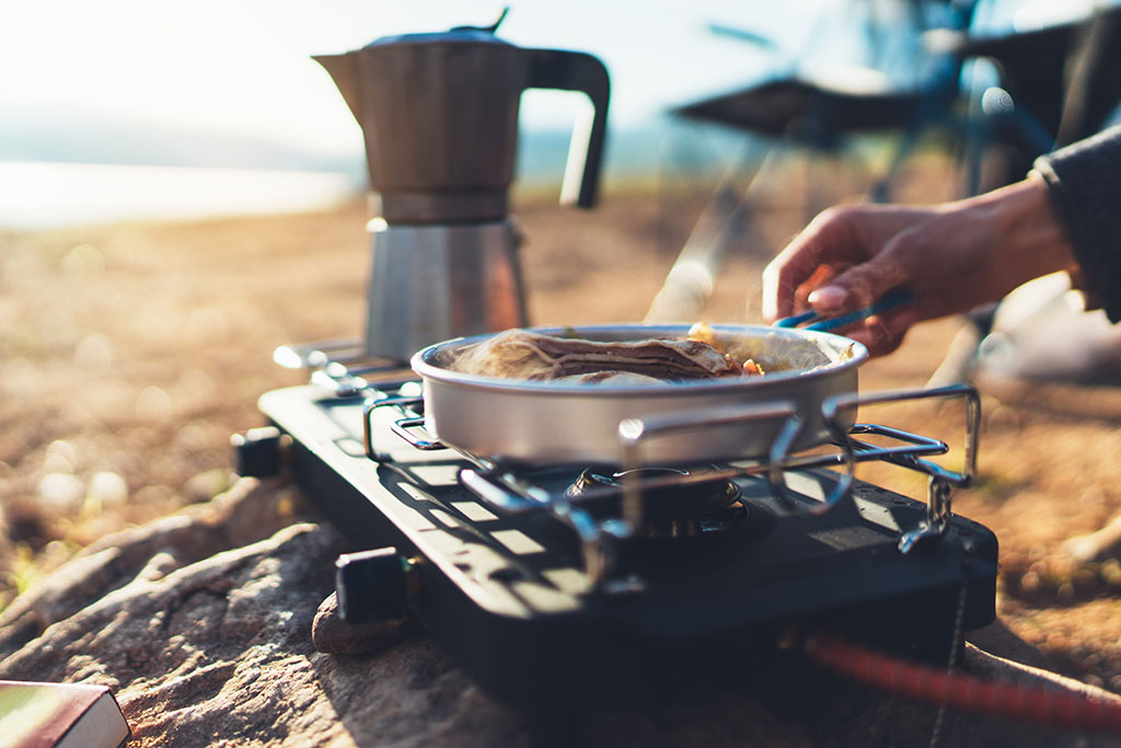 someone sitting outside in the countryside cooking in a pan on a camping stove with a pot of coffee in the background.