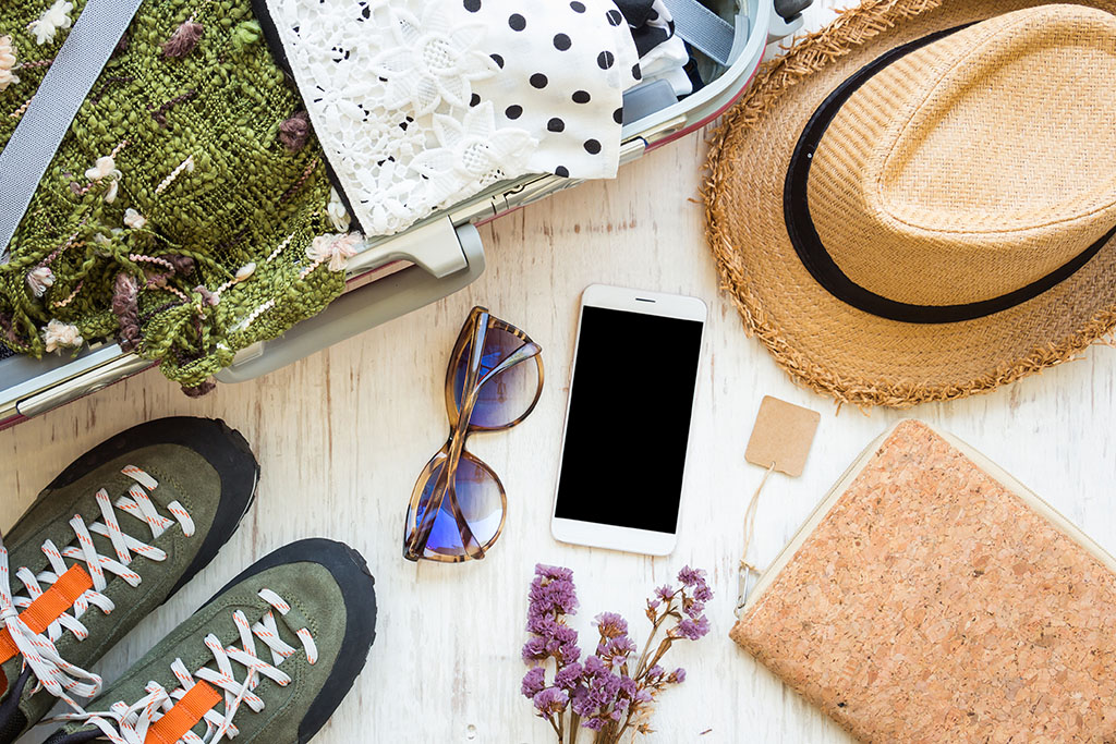 various travel items including a straw hat, trainers, sunglasses and a mobile phone next to open luggage on a white background.