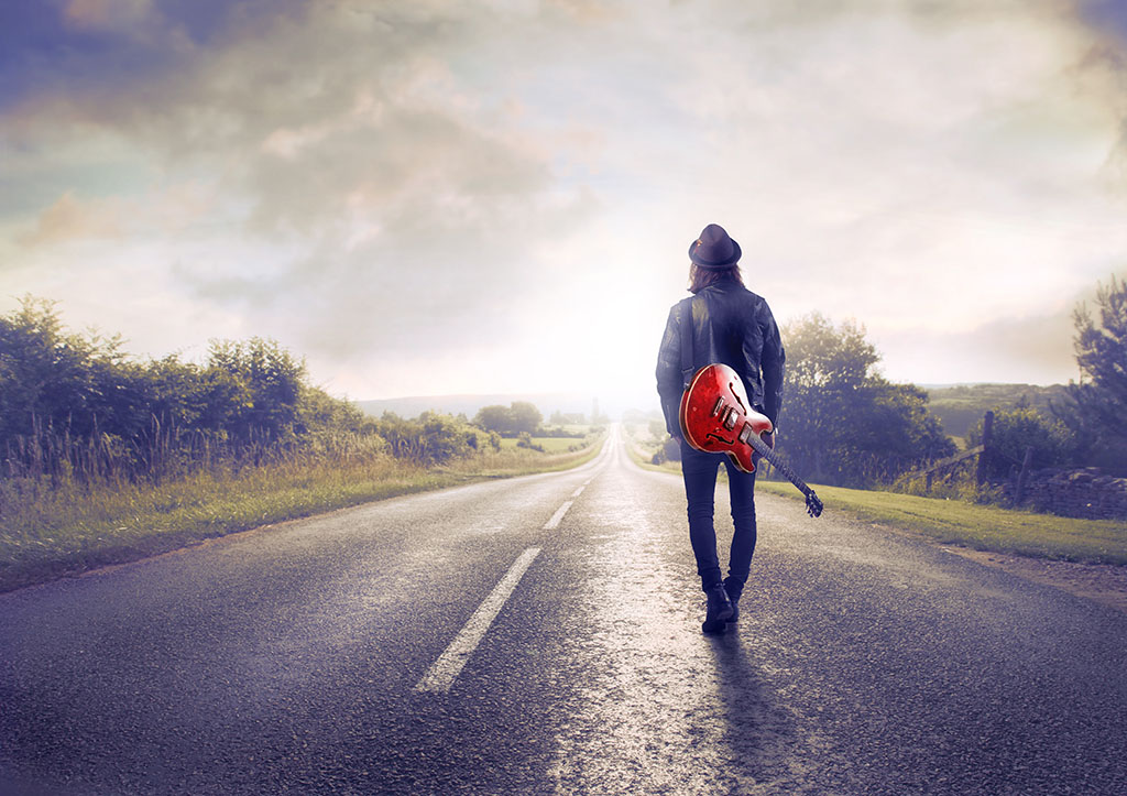 Young musician walking on a countryside road with a guitar on his shoulder