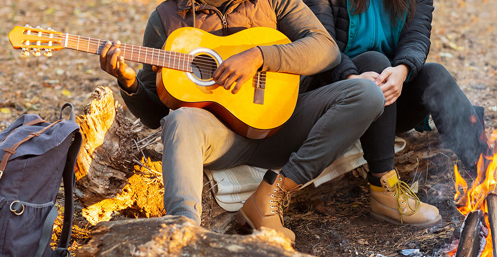 Cropped of mixed race couple enjoying time at forest, camping, playing guitar next to bonfire, panorama