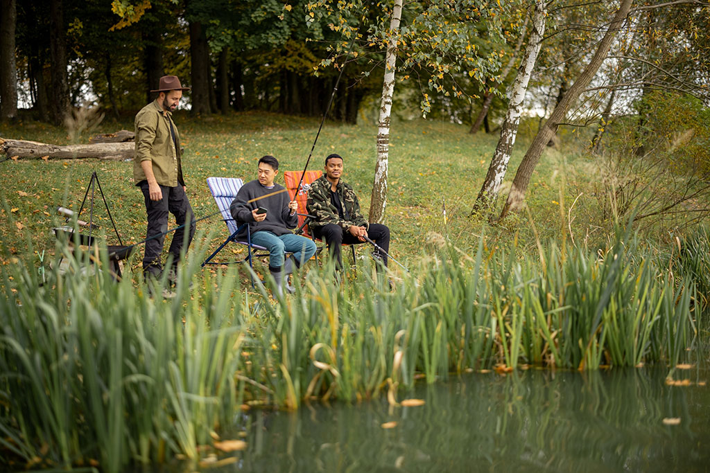 Pleased multiracial male friends resting and fishing on river or lake coast. Concept of leisure, hobby and weekend in nature. Idea of friendship and spending time together. View from the waterside